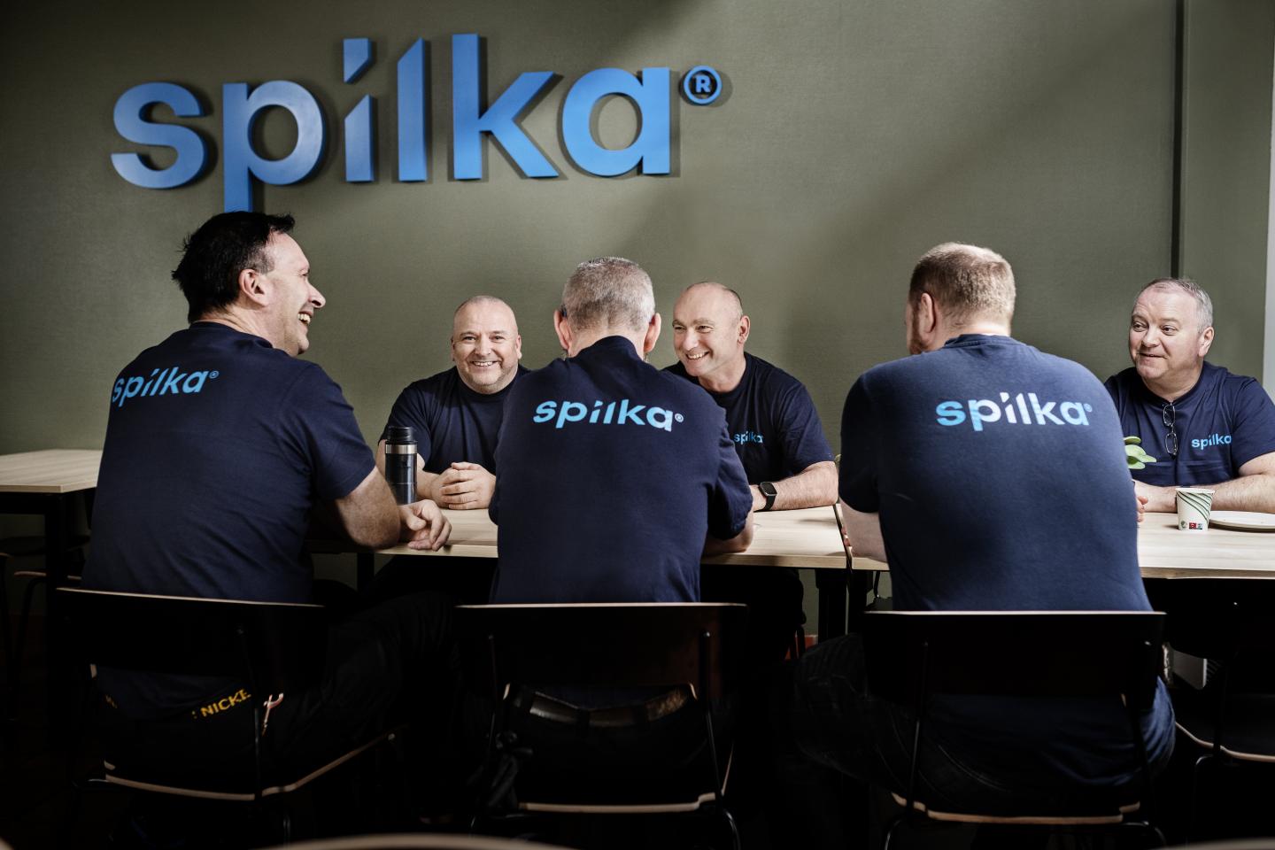 Six men in navy "spilka" shirts sit at a table, smiling and engaging in conversation in front of a large "spilka" sign on the wall.