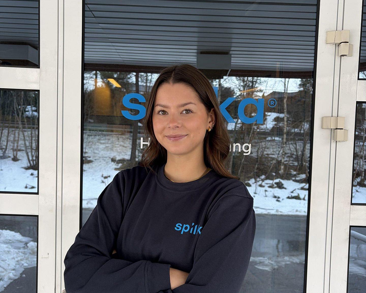 A young woman with brown hair and a black sweatshirt stands confidently with arms crossed outside a building with snow and trees in the background.
