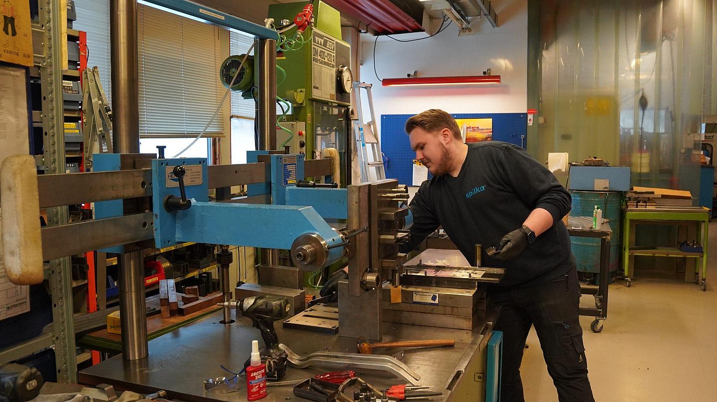 Mann jobber med maskin i verksted med verktøyhylle, arbeidsbord og skap i bakgrunnen.