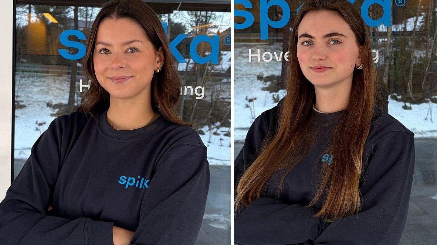 Two women with long brown hair and crossed arms stand in front of a glass window with blue "Spika" branding, outdoors on a snowy day.