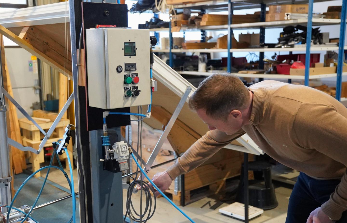 A man inspecting or adjusting machinery in a workshop with shelves of tools and supplies, focusing on a control panel and connected equipment.
