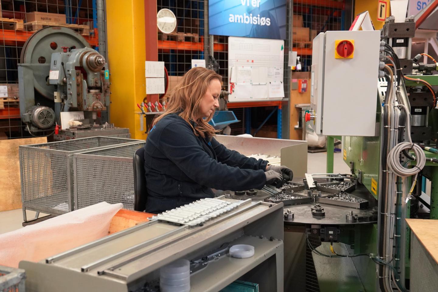 A woman working with machinery on an assembly line in an industrial setting. She wears gloves and concentrates on assembling parts amid technical equipment.