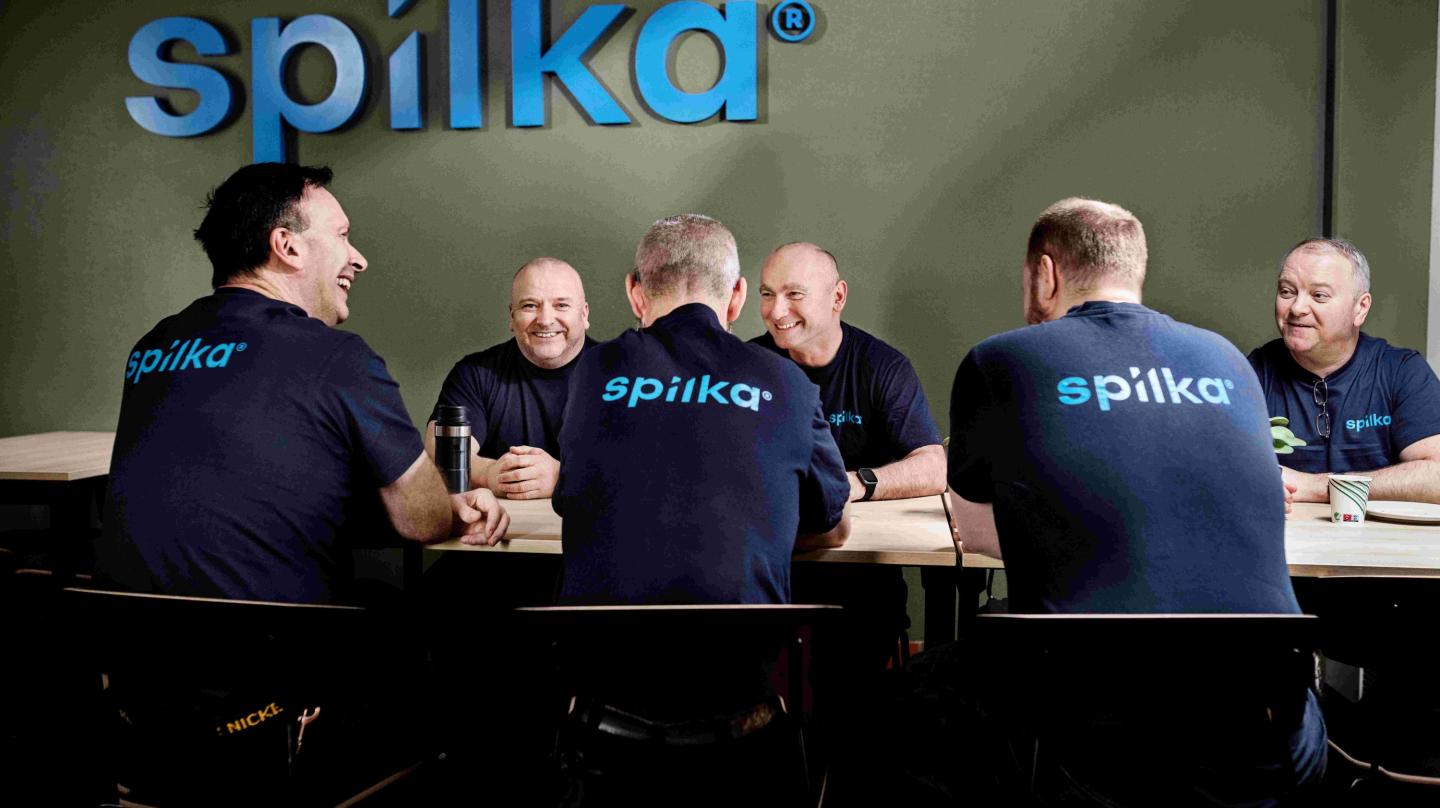 Syv personer i blå T-skjorter med "spilka" på, sitter rundt et bord og ler sammen i et møterom med grønn vegg og stort "spilka" logo.