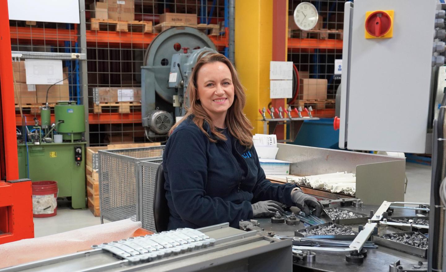 A woman with brown hair working at a machine in an industrial warehouse, surrounded by equipment, tools, and storage shelves.