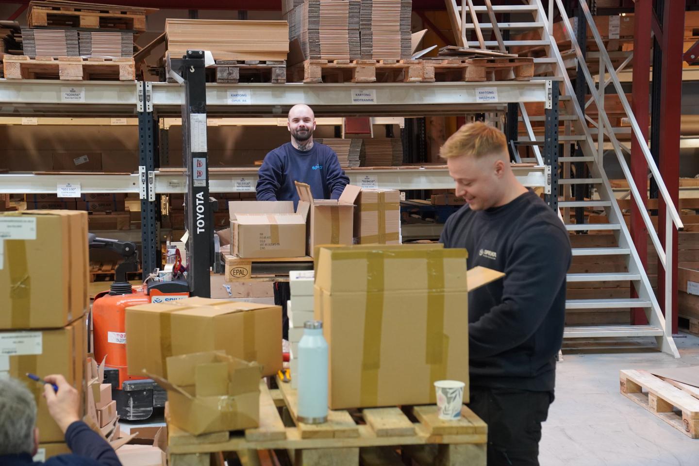 To arbeidere pakker esker i lager, med paller og hyller med pakket gods og tretrapper i bakgrunnen.