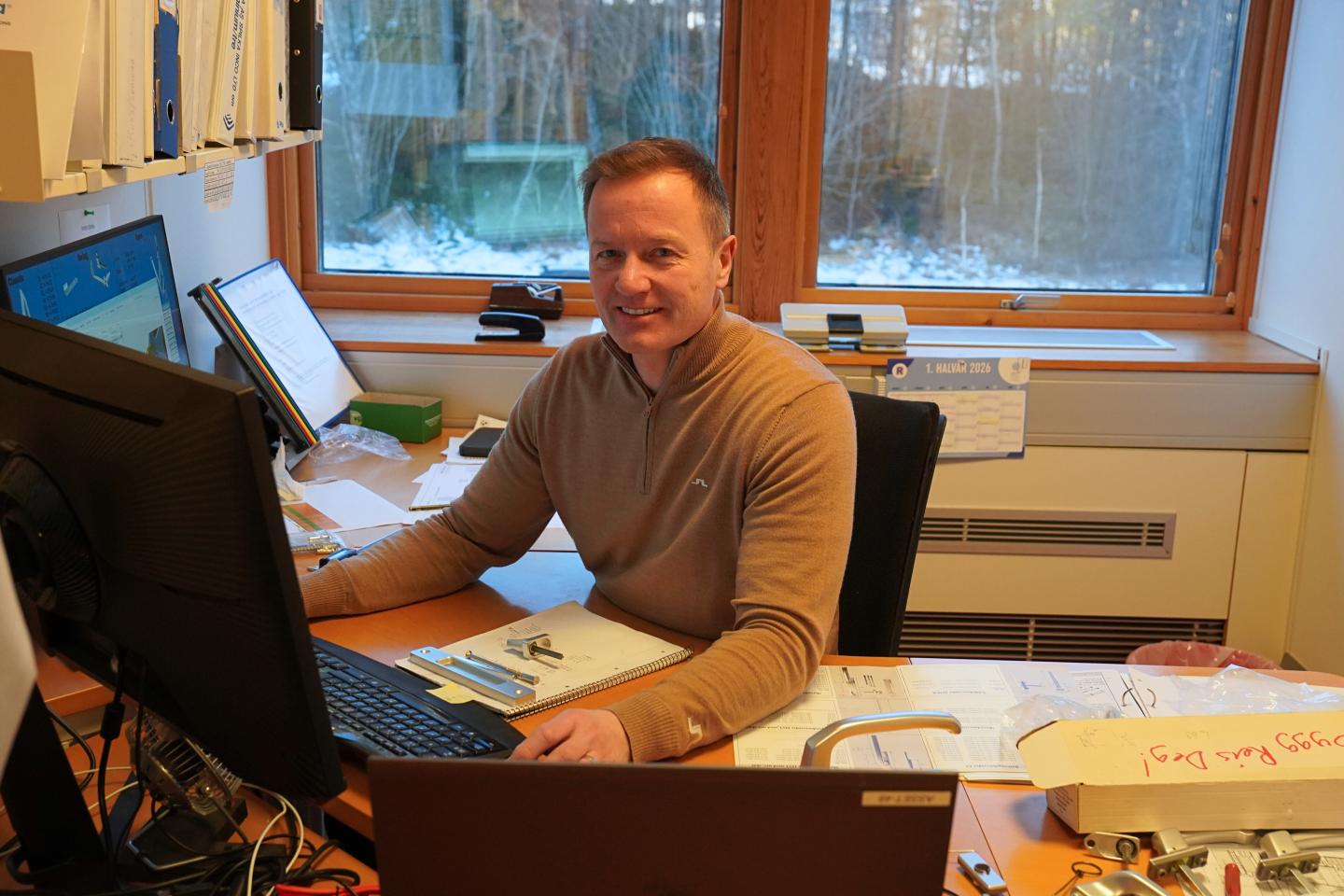 En mann sitter ved et skrivebord med to dataskjermer, omgitt av papirer, kontorrekvisita og en kalender foran et vindu med vinterutsikt.