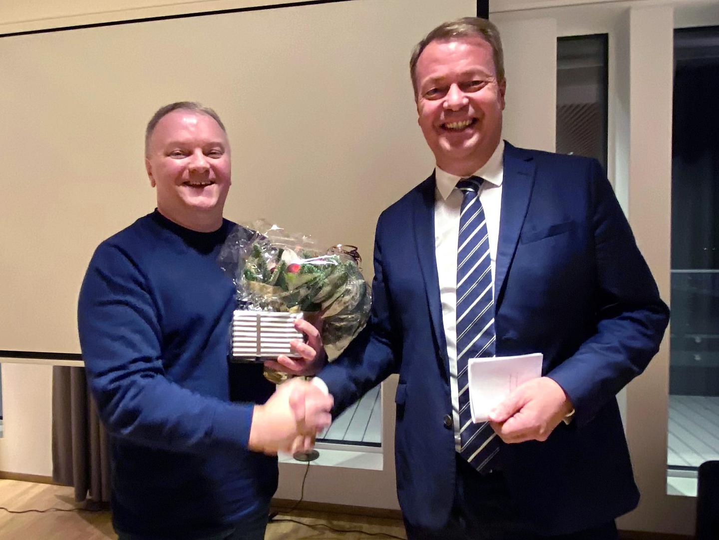 To personene veksler håndtrykk, én holder en bukett blomster og den andre et stykke papir, begge smiler under en formell anledning.