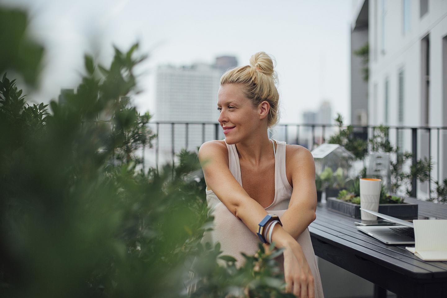 A woman with blonde hair in a bun relaxes on a balcony, surrounded by plants, with a cityscape and tall buildings in the background.