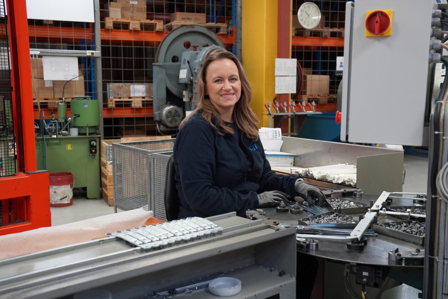 Kvinne i arbeidsdrakt og hansker smiler mens hun jobber med maskindeler i et industrilaboratorium eller verksted.