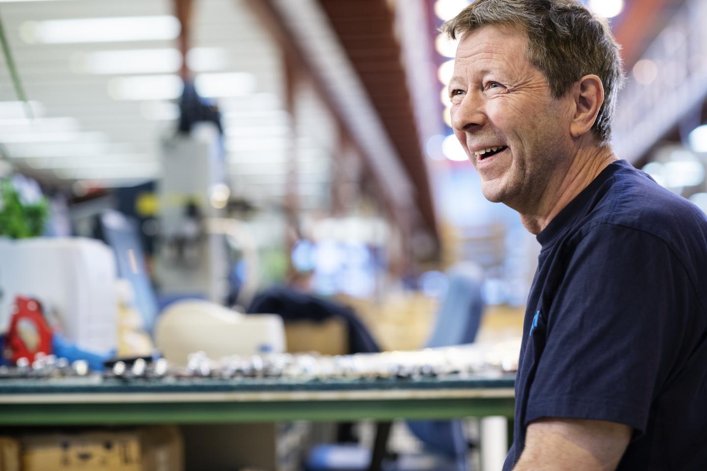 En eldre mann med kort hår smiler mens han ser mot høyre i et verksted eller fabrikkmiljø. Bakgrunnen er uklar med ulike maskiner og utstyr.