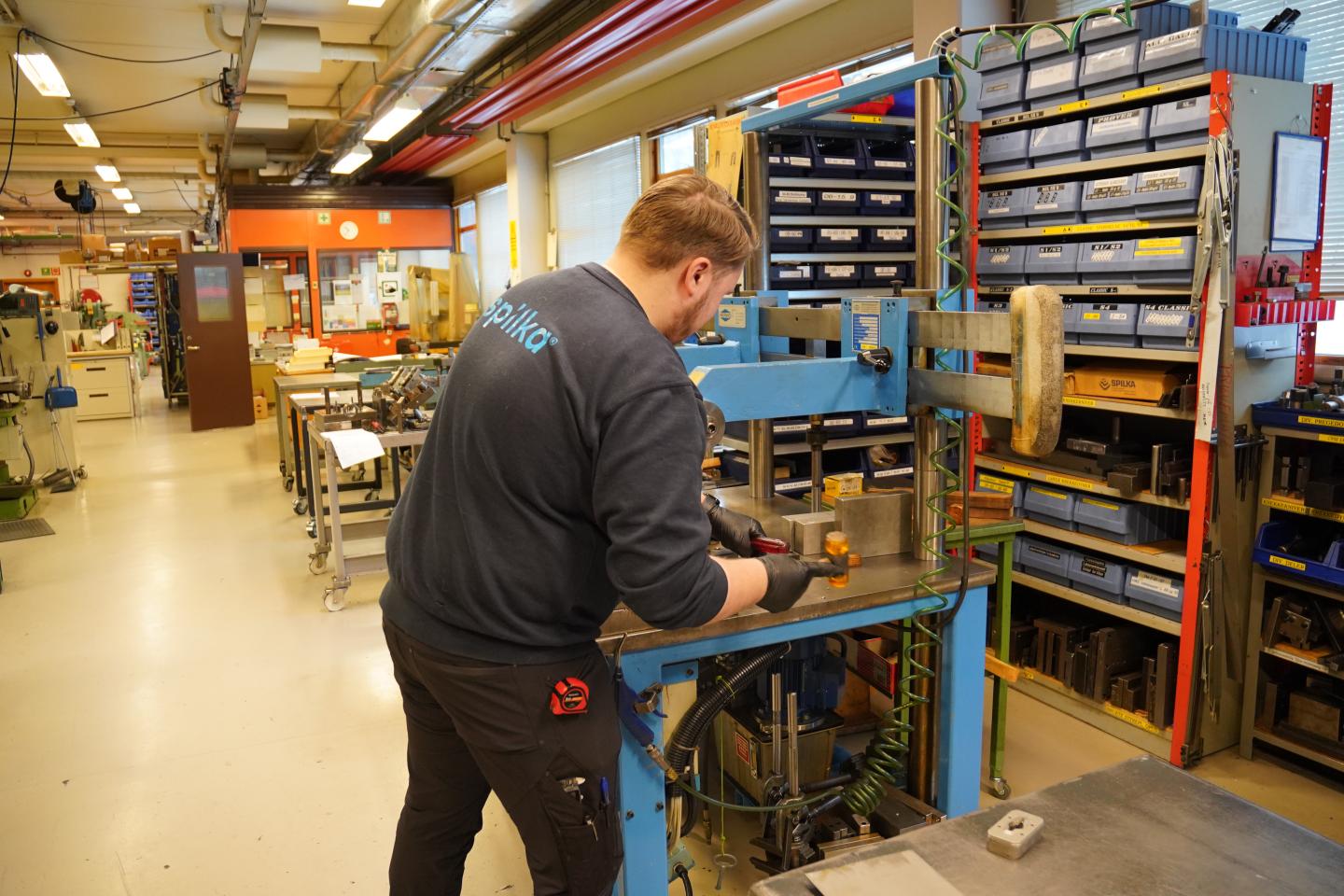 A man working on a metal machine in a well-organized workshop with storage shelves, tools, and equipment in the background.