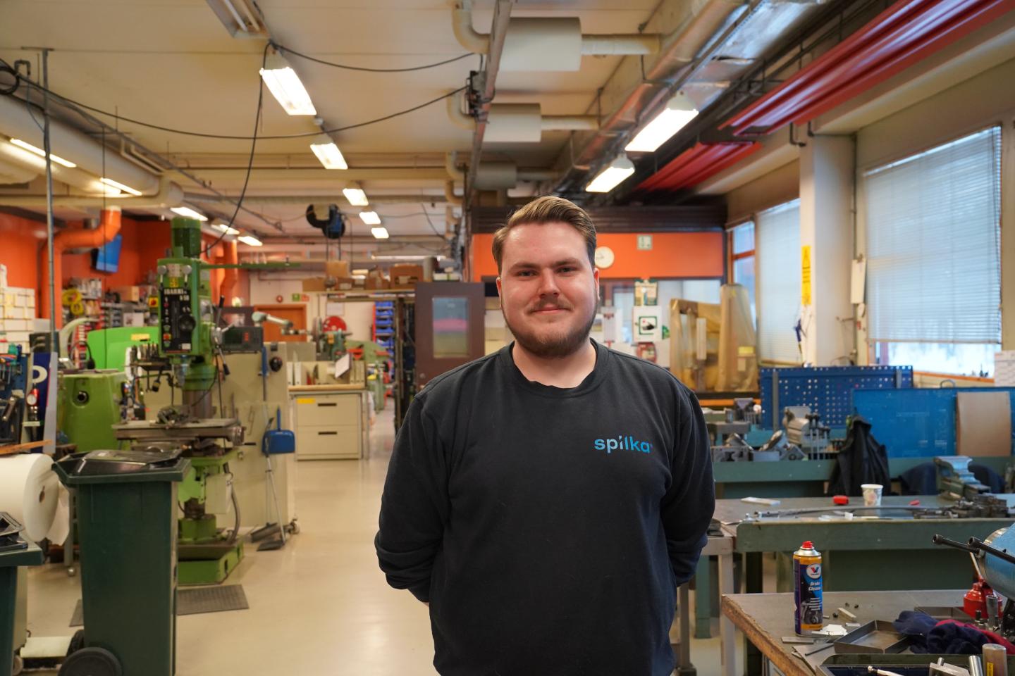 A man with short hair and a beard stands in a workshop filled with various machinery and tools, wearing a black shirt with "spilka" logo.
