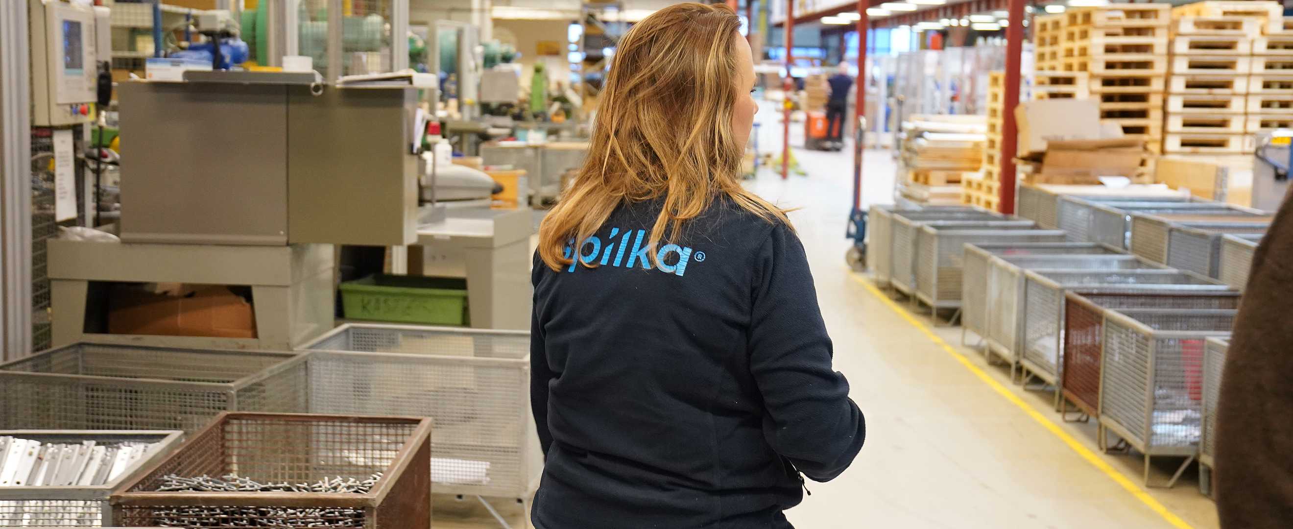 A woman with long blonde hair wearing a black jacket with "Voilka" on the back, standing in a warehouse or industrial setting with storage bins and pallets.